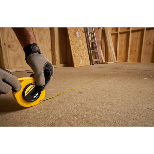 STANLEY 0-34-108 measuring tape, yellow, used on construction floor, person wearing gloves.