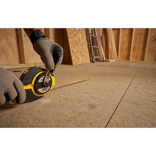 Photo of STANLEY 0-34-134 long tape measure in use on a construction floor, gloved hands holding the tool, side angle.
