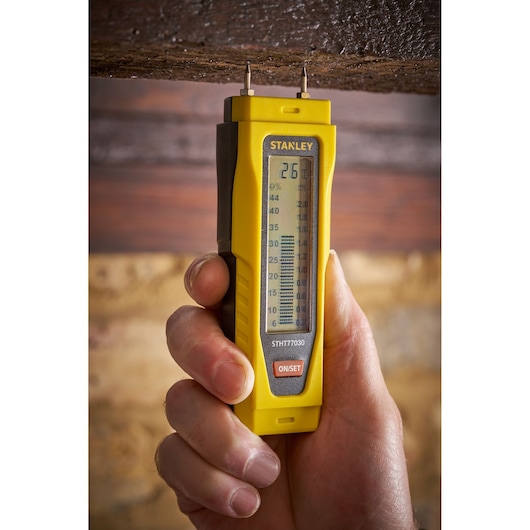 STANLEY 0-77-030 moisture meter held to wood, yellow handheld tool, front view.