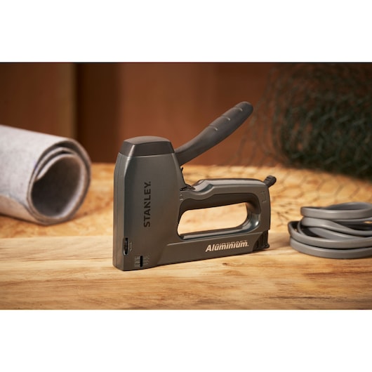 STANLEY 0-TR250 aluminum staple gun on a wooden surface, side view, with rolled materials nearby.