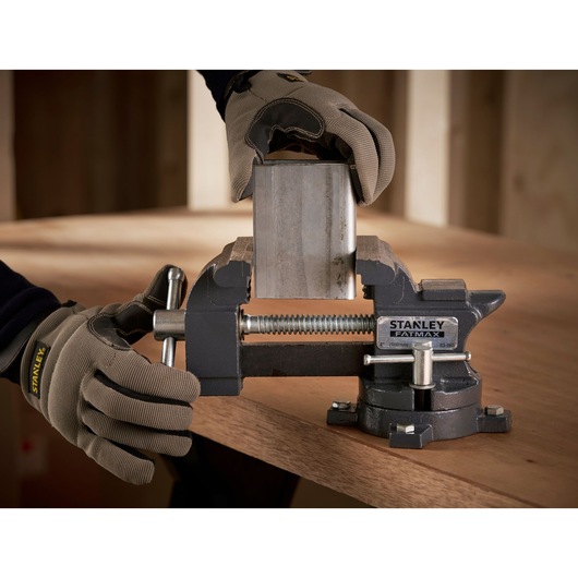 Photo of STANLEY 1-83-065 bench vise clamping a metal piece on a workbench, worker wearing gloves.