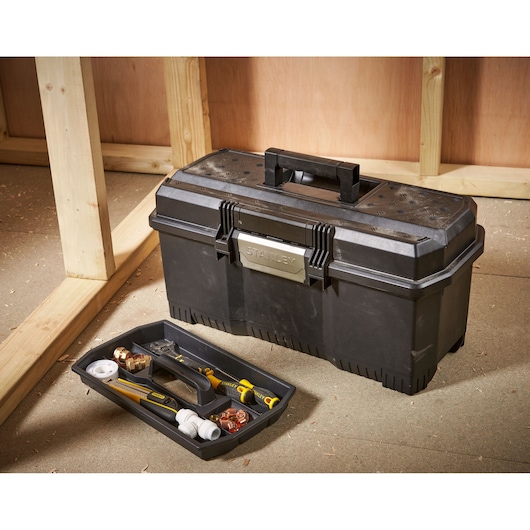 A black Stanley toolbox with a handle and latch, placed on a workshop floor. The toolbox tray is shown in front, containing several hand tools and plumbing fittings.
