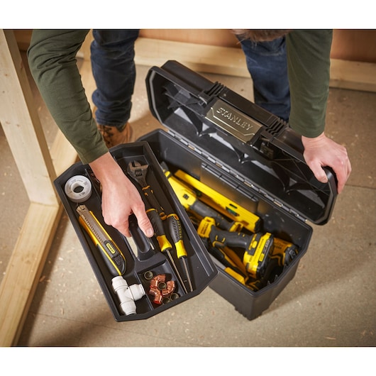 A person opens a black STANLEY toolbox containing various hand tools such as screwdrivers, a wrench, a utility knife, tape, and fittings. The toolbox has different compartments for organization and contains yellow and black tools.