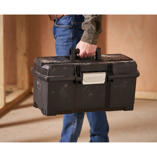 Close-up of a person holding a black STANLEY toolbox by its handle in a workshop setting.