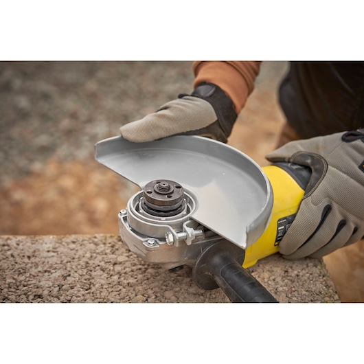 STANLEY FMEG232 angle grinder close-up, yellow body, safety guard adjustment, glove hands, outdoor setting.