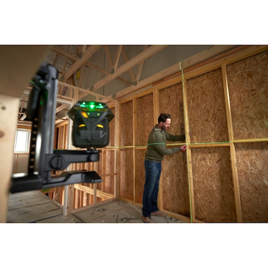 STANLEY FMHT1-77356 green laser level projecting horizontal and vertical lines in wood-framed room, worker marking wall.