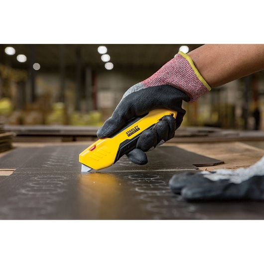 Stanley FMHT10369-0 utility knife cutting material, held by gloved hand in industrial setting.