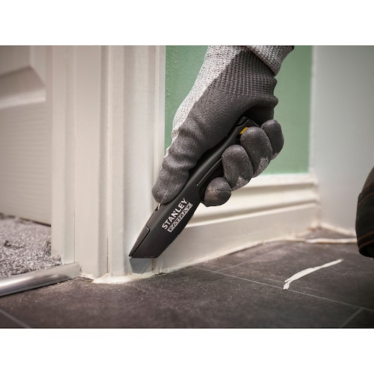 Photo of STANLEY FMHT10500-0 utility knife cutting caulk at door frame corner on black tile floor.