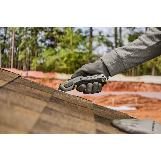 A gloved hand is using a Stanley utility knife to cut roofing shingles on a rooftop, with trees and construction materials in the background.