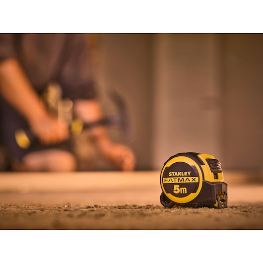 STANLEY FMHT33100-0 tape measure FATMAX 5m in focus, construction setting with blurred figure in background.