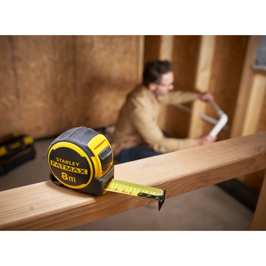 Photo of STANLEY FMHT33102-0 FATMAX 8m tape measure on wood beam with worker in background.