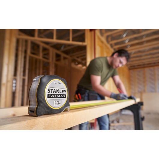 STANLEY FATMAX 10m tape measure FMHT38232-0 on wood beam, construction worker measuring in background.