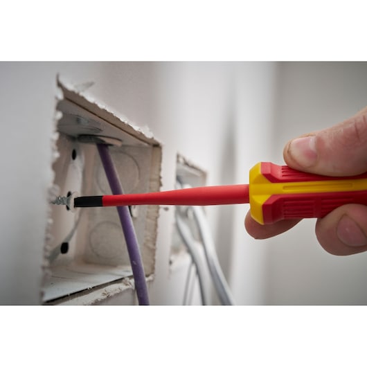 Insulated screwdriver fastening a screw inside an electrical box with visible wires in a wall opening.