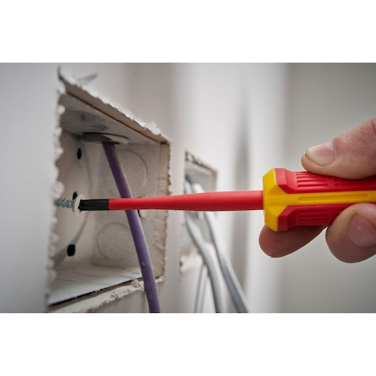 A red and yellow insulated screwdriver is used to tighten a screw inside an electrical wall box.