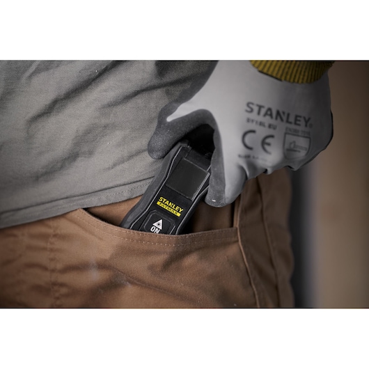 Photo of STANLEY FMHT77165-0 laser distance measurer in gloved hand, being placed in brown pants pocket.