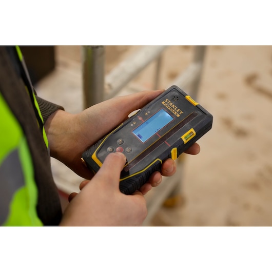 Photo of STANLEY FMHT77652-0 digital detector held in hand, construction site background visible.