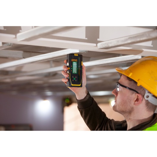 STANLEY FMHT77653-0 laser level held by worker in yellow helmet measuring metal ceiling frame.