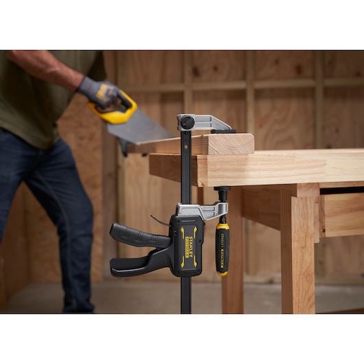 STANLEY FMHT83008-0 clamp securing wood on workbench, person sawing wood in workshop.