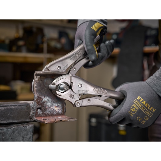 Stanley locking pliers grip a metal piece while hands in Stanley gloves hold the tool in a workshop setting.