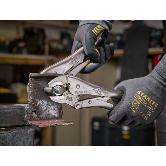 Stanley locking pliers held by gloved hands gripping a metal piece in a workshop setting.