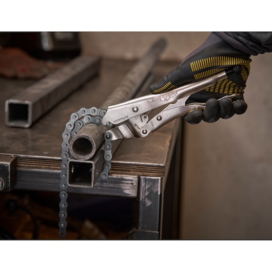 A gloved hand uses a Stanley chain locking plier to grip a round metal pipe on a workbench.
