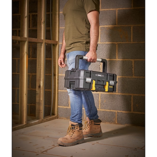 STANLEY FMST1-71967 toolbox with black and yellow latches, carried by person in work boots at construction site.