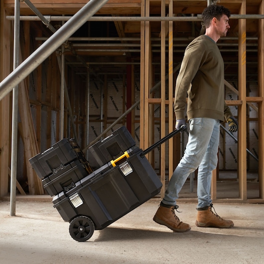 STANLEY FMST17870-1 rolling toolbox with black and yellow accents, stacked with toolboxes, pulled by worker on construction site.