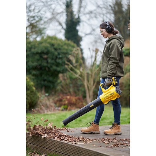 Photo of STANLEY SFMCBL7B cordless leaf blower in use outdoors, yellow and black, clearing leaves from wooden deck.