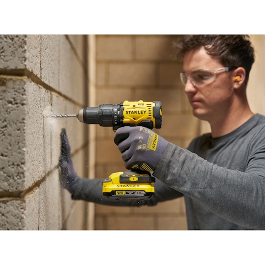 Person drilling into a concrete wall with a yellow STANLEY cordless drill, wearing gloves and safety glasses.