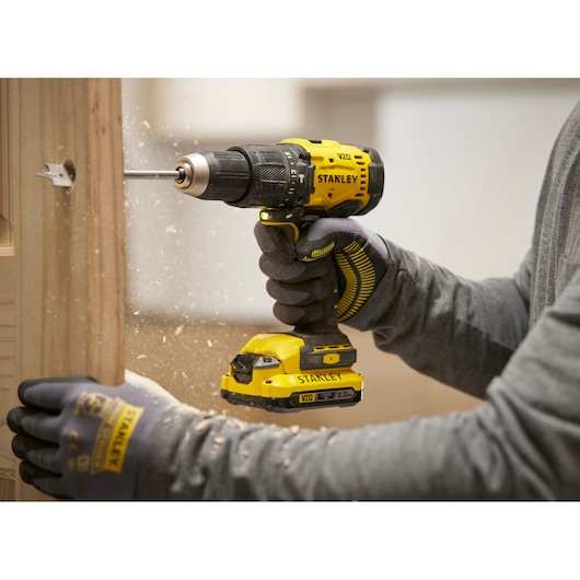Person uses a Stanley V20 cordless drill to bore a hole in wood, wearing protective gloves and creating wood shavings.