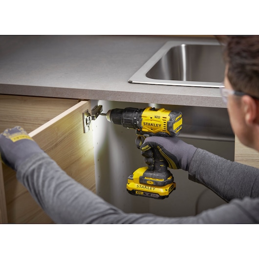 Person uses STANLEY V20 drill on a cabinet hinge under a kitchen countertop near sink.