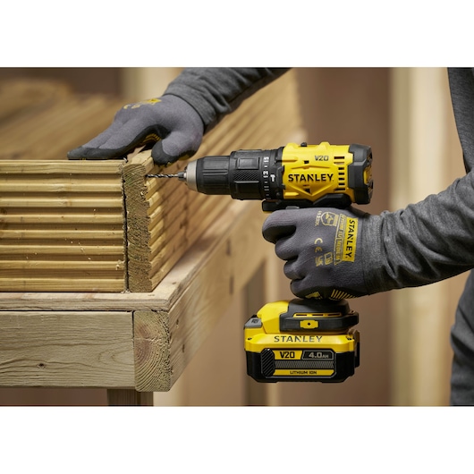 Person using a Stanley V20 cordless drill to drill into stacked wooden planks, wearing gloves.