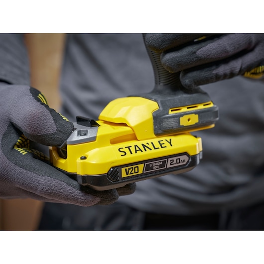 Close-up of gloved hands holding a yellow STANLEY V20 2.0Ah lithium-ion power tool battery.