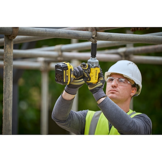 Worker uses STANLEY cordless impact driver SFMCF921B on steel scaffolding outdoors, wearing gloves and safety gear.