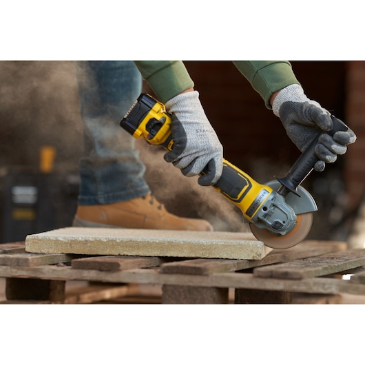 STANLEY SFMCG700B angle grinder cutting stone slab on pallet, worker wearing gloves and boots.
