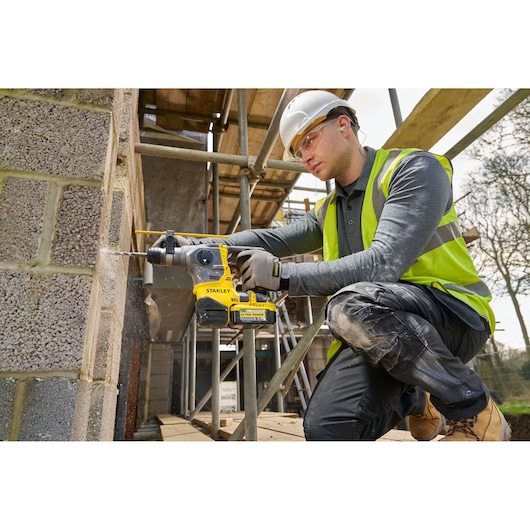 Person drills wall using a Stanley V20 rotary hammer at a construction site, wearing safety gear.