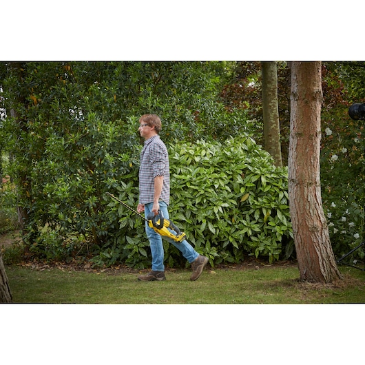Photo of STANLEY SFMCHT855B hedge trimmer held by person walking outdoors near greenery.