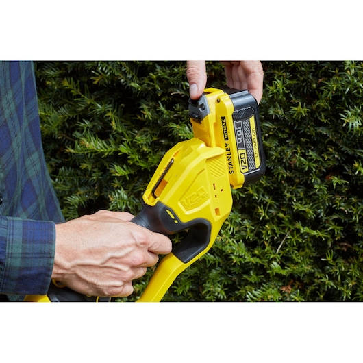 STANLEY SFMCPH845M1 hedge trimmer handle with 18V battery shown outdoors near green bushes.