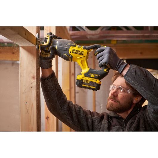 STANLEY SFMCS311M1T cordless reciprocating saw in use cutting wooden beam, construction site background.