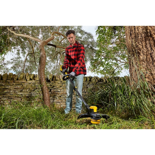 STANLEY SFMCSTB933M string trimmer used for grass trimming in a garden with stone wall and trees.