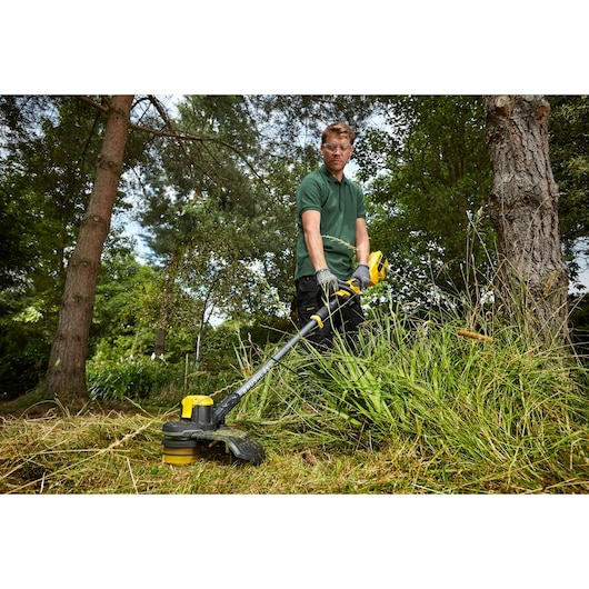 STANLEY SFMCSTB963B grass trimmer in use outdoors cutting tall grass, handled by a person in gardening gloves.