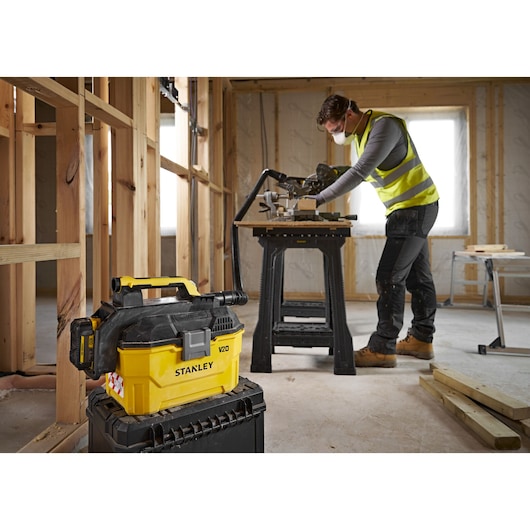 Stanley V20 vacuum in foreground on a toolbox, worker in safety vest using saw in unfinished room.