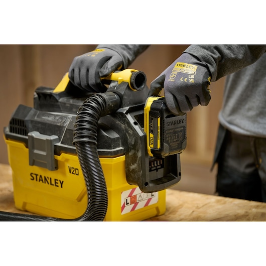Stanley V20 cordless vacuum in use, person with gloves installing battery on yellow and black unit.