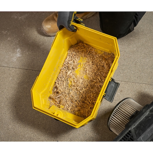 Yellow vacuum container filled with wood shavings, shown on a workshop floor with a filter and a gloved hand.