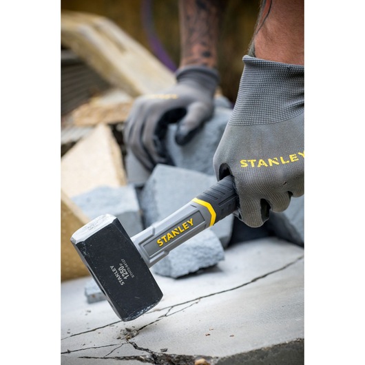 Photo of STANLEY STHT0-54127 club hammer in use, black head with yellow accents, held by gloved hand on concrete.