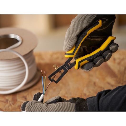 STANLEY STHT0-75068 wire stripper held by gloved hand, stripping colored wires on wooden surface.
