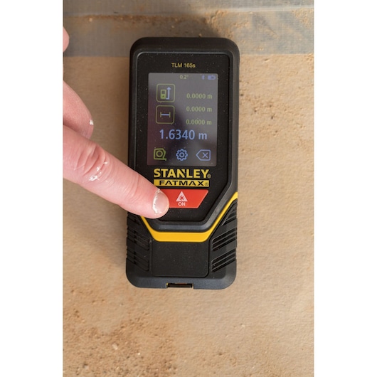 Photo of a STANLEY STHT1-77139 laser distance measurer in use, finger pressing power button, close-up view.