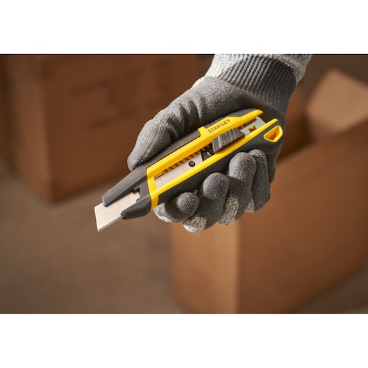 STANLEY STHT10501-0 utility knife held in gloved hand, yellow and black body, cardboard boxes in background.