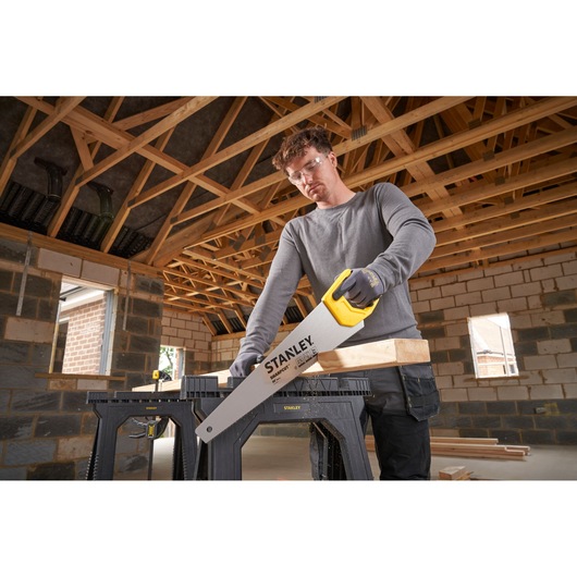 Photo of STANLEY STHT20367-1 hand saw with yellow handle, cutting wood on a sawhorse in a building under construction.