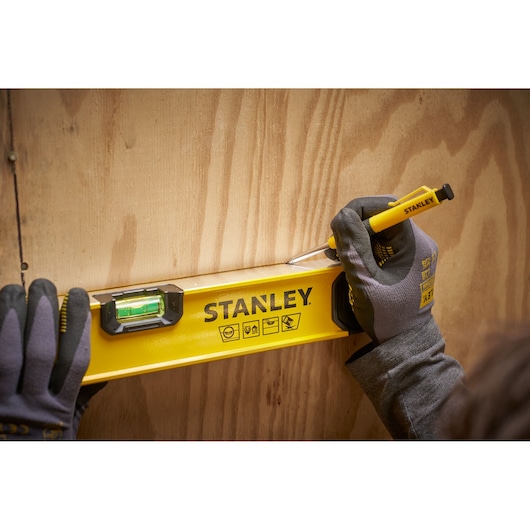Close-up of gloved hands using a yellow STANLEY spirit level against a wooden surface, while marking a line with a yellow STANLEY pencil.